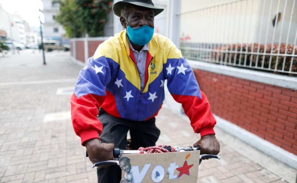 A pro-government supporter dressed in a Venezuelan flag jacket cycles through the city with a sign that reads "VOTA"