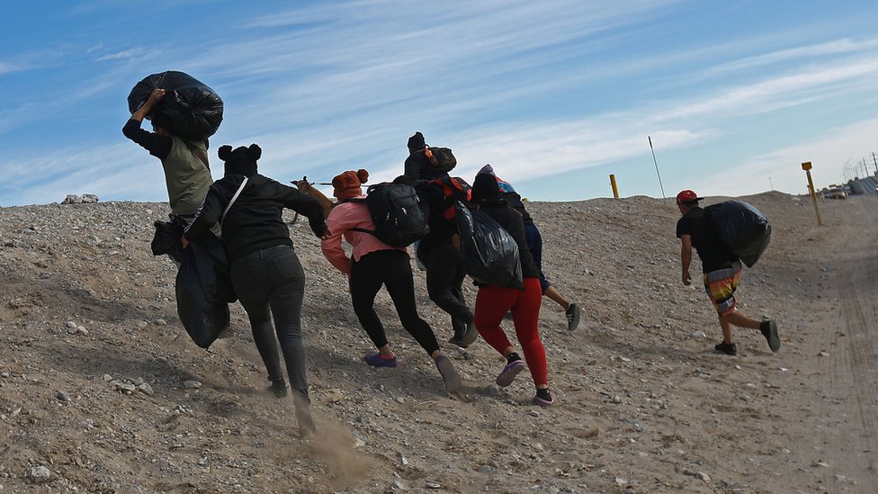 A group of people with their backs turned walking up a hill to try and cross the US Mexico border