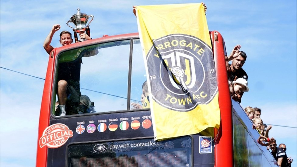 Джош Фолкингем из Харрогейт-тауна держит трофей, когда празднует с товарищами по команде во время автобусного тура с открытым верхом вокруг Харрогейта