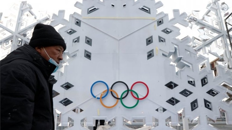A man wearing a mask and coat walks past a building bearing the Olympics logo