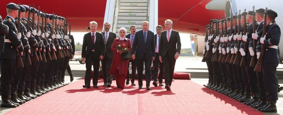 Г-на Эрдогана и его жену вместе с высокопоставленными официальными лицами сопровождают по красной ковровой дорожке, окруженной военным почетным караулом.
