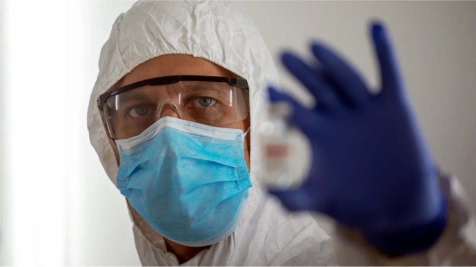 A man in a hazard suit holds a vaccine vial