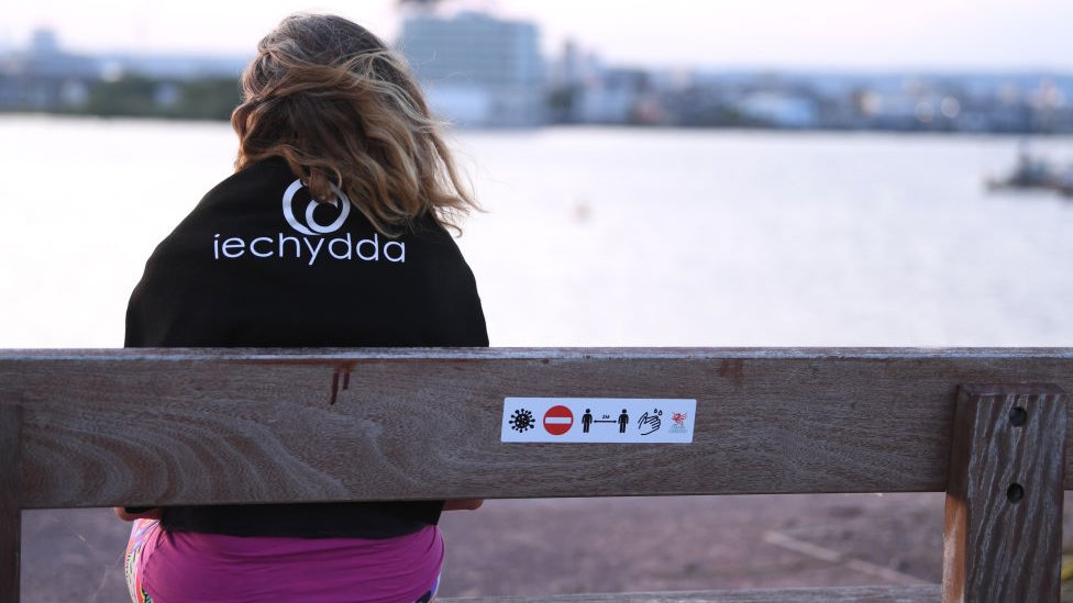 Женщина сидела на скамейке в парке в Кардиффе с предупреждающим знаком на спине