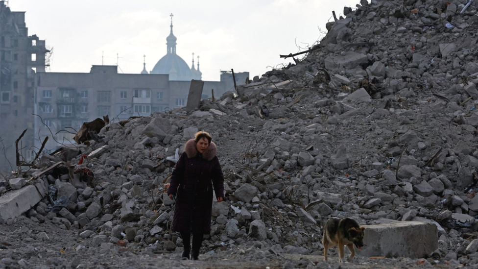 A woman and a dog walk in the rubble of Mariupol