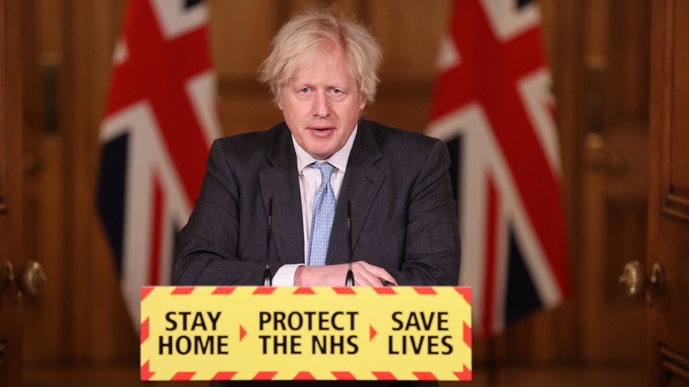 Boris Johnson standing at a government press briefing podium with the slogans 'Stay home, protect the NHS, save lives' printed on the front