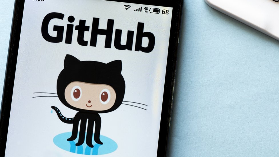 Здесь изображен логотип Github - гибрид осьминога и кошки с почти человеческим лицом в мультяшном стиле. Это видно на телефоне на фоне голубого стола возле ноутбука