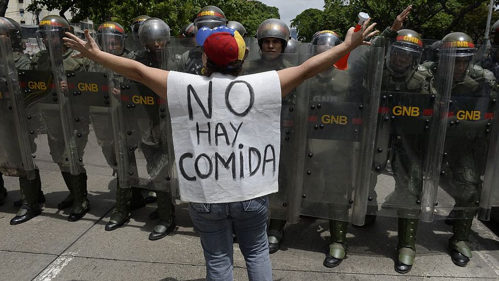 El Fondo Monetario Internacional estima que la inflación en Venezuela a final de año será del 720%. Mujer con un chaleco que dice: "No hay comida", parada frente a policías venezolanos.
