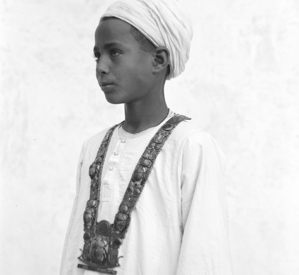 Foto em preto e branco mostra um menino egípcio de semblante sério com roupas brancas e um turbante usando um colar longo e cheio de joias, o pendente principal forma um escaravelho