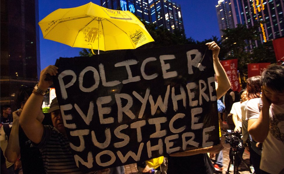 Протестующие, выступающие за демократию, держат желтый зонтик - символ продемократического движения территории - и баннер с надписью «полиция везде, правосудие нигде», недалеко от места, где Чжан Дэцзян выступил с программной речью 18 мая