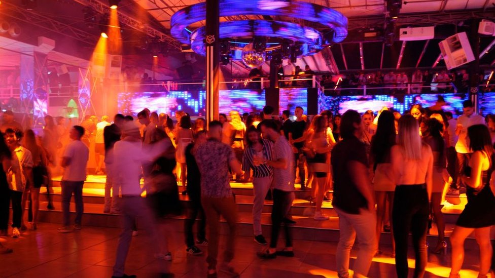 Jovens dançando em um clube onde há imagens coloridas em telões nas paredes