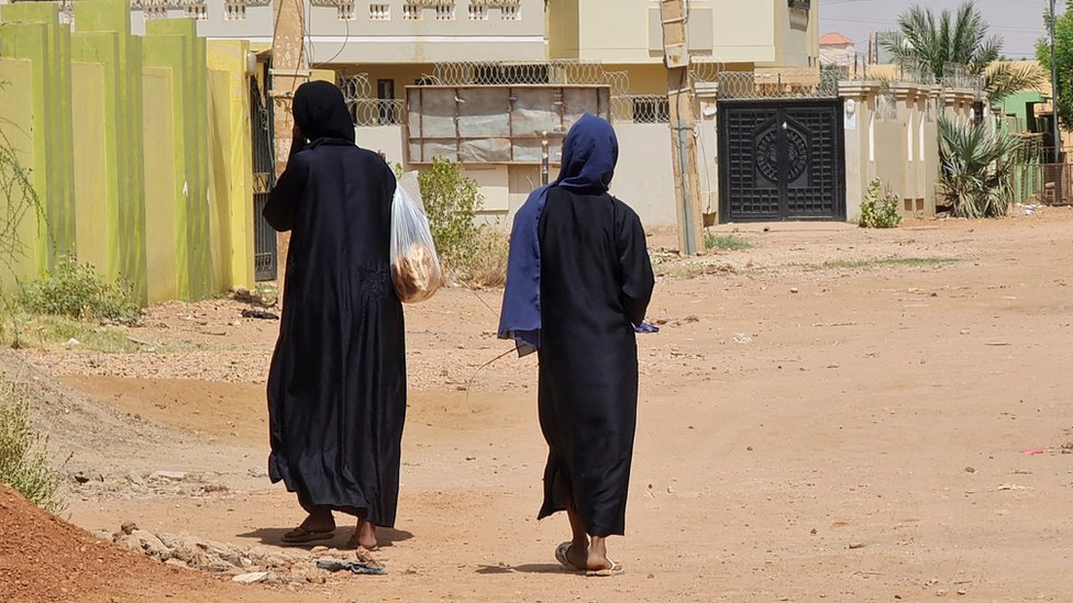 Two women in black cloaks walking towards residential buildings, their backs to the camera. One woman is carrying a bag full of bread.