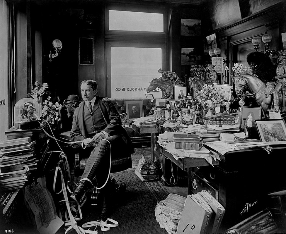 Corretor de ações Thomas William Lawson sentado em frente a uma máquina, em meio a livros e outros objetos em seu escritório