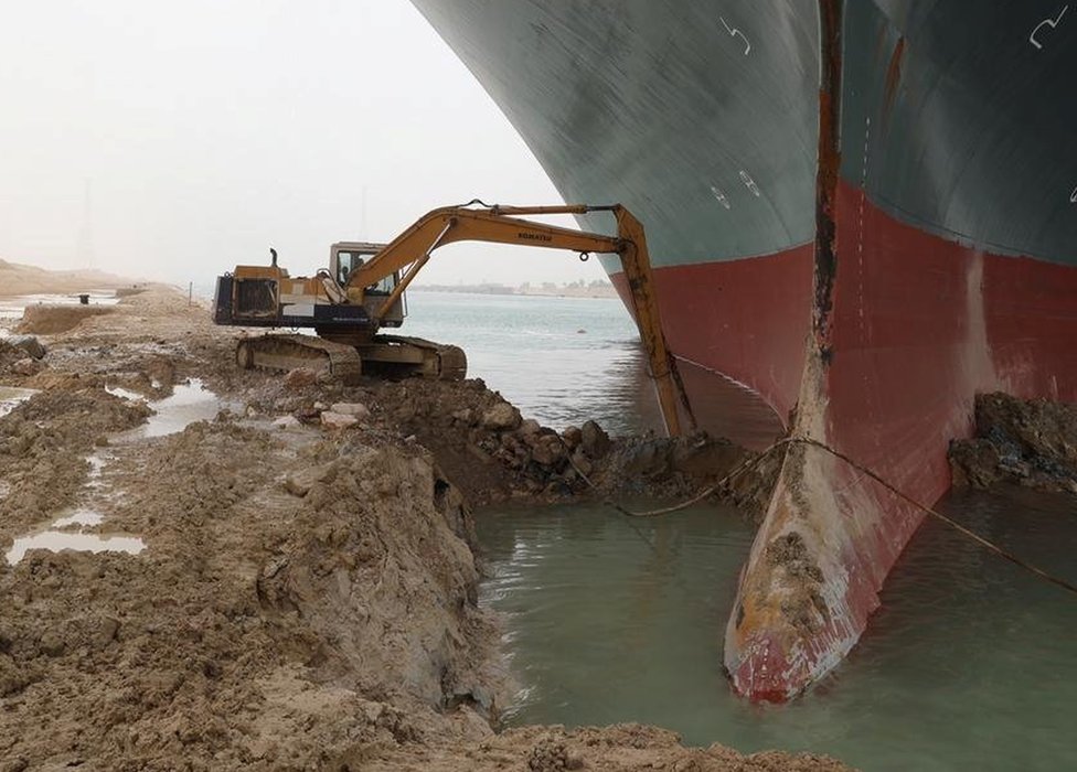 Una excavadora en tierra intenta apartar la arena y el lodo alrededor del caso del barco.