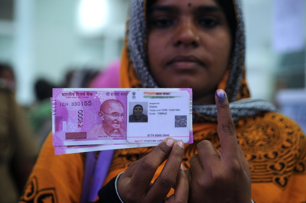 Индийская женщина позирует с новыми банкнотами в 2000 рупий, своим удостоверением личности Aadhaar и пальцем, исписанным несмываемыми чернилами, после обмена изъятых банкнот 500 и 1000 рупий в банке в Ченнаи 17 ноября 2016 года