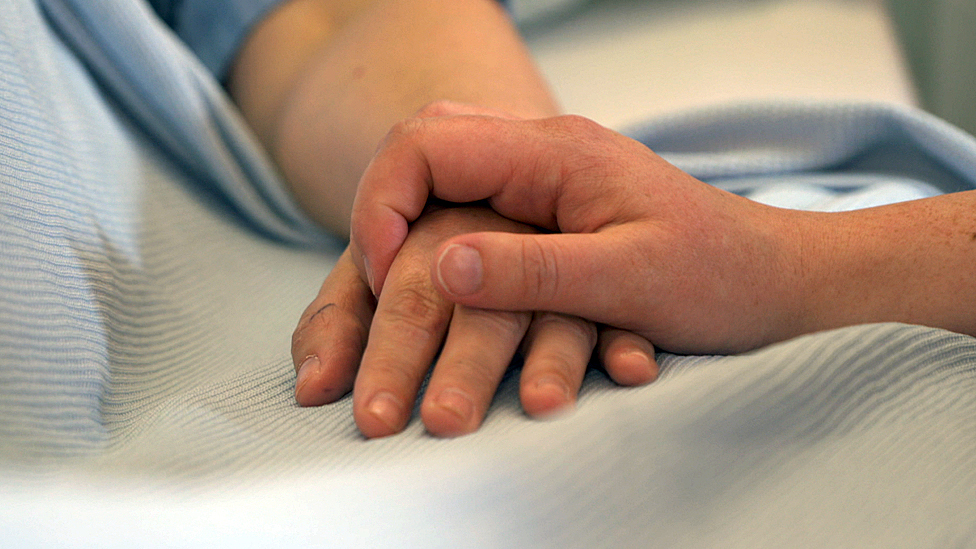 A hospital patient's hand is held by the hand of another person