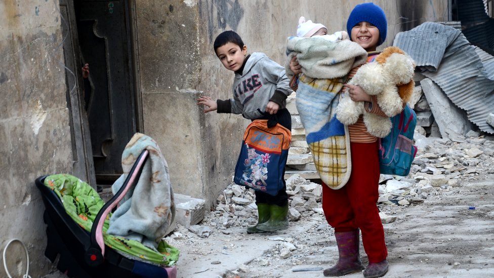 Сирийская девочка, держащая своего младшего брата, стоит рядом с развалинами