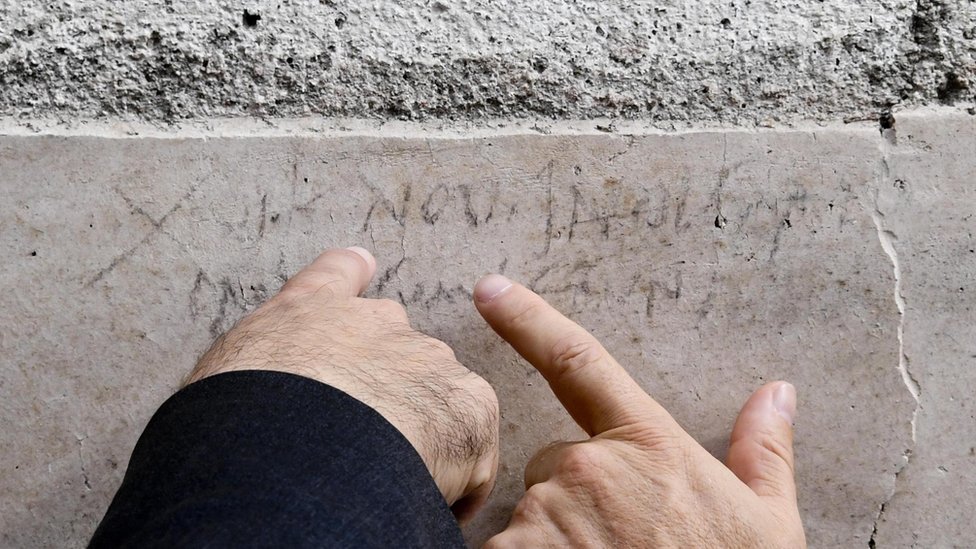 Руки двух мужчин указывают на выцветшую надпись на каменной кладке стены. Буквы NOV, в основном непонятные для тех, кто не читает древнюю латынь, хорошо видны - отсылка к ноябрю