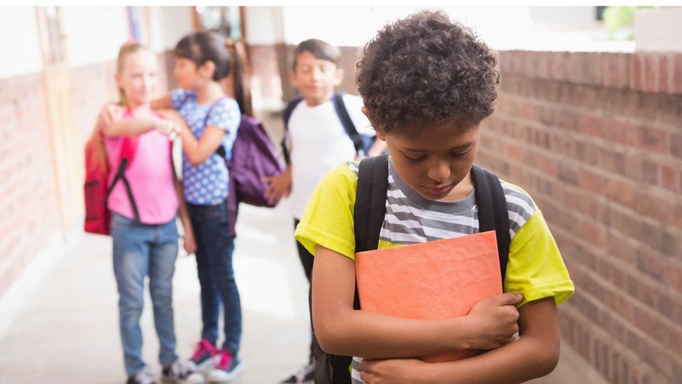 A boy in the foreground bows his head, a girl behind him points, a girl has her arms around the girl as if the placate her and a boy stands near them.