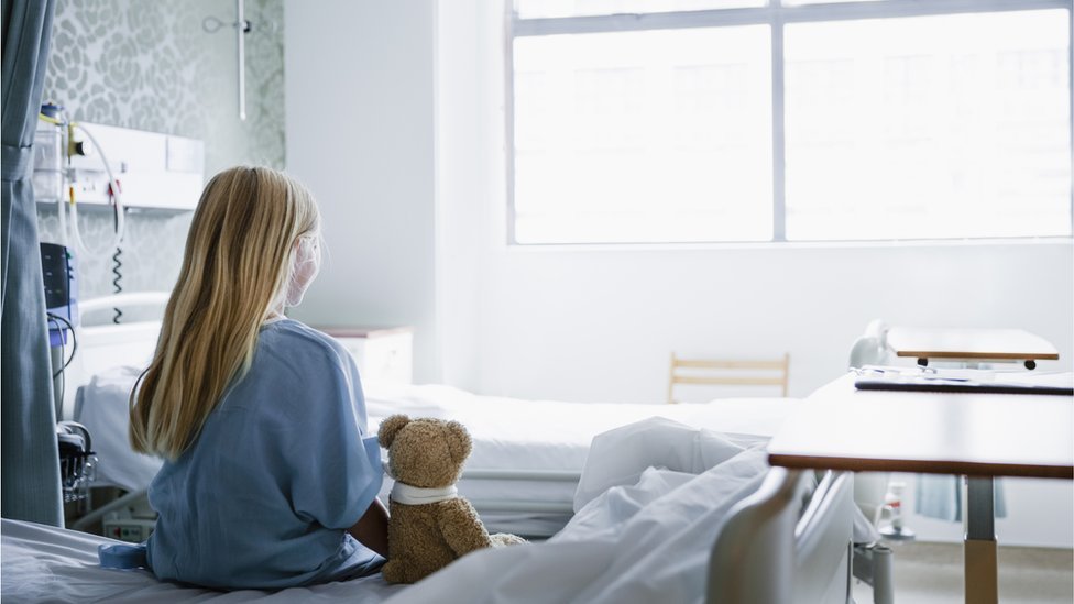 Young girl sitting on a hospital bed with a teddy bear next to her