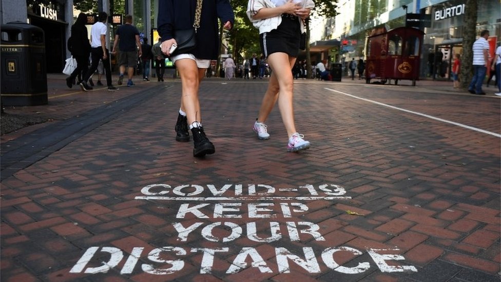 People walking past a pavement sign reading "Covid-19 - keep your distance"