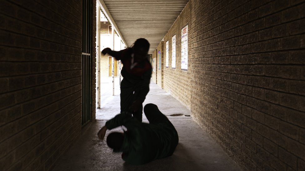 Silhouettes of a child towering over another child on the ground in a dark alleyway, with the child that is standing about to hit the child on the ground