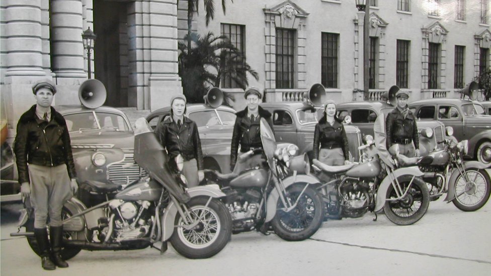 Fotografia em preto e branco mostra jovens vestidos com jaquetas de couro ao lado de carros e motos