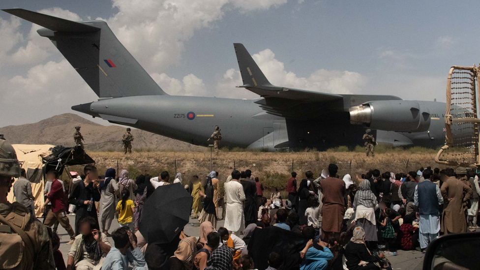 Personas haciendo cola en el aeropuerto cerca de un avión rodeado de tropas.