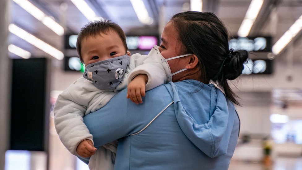 Bebê no colo da mãe, ambos de máscara contra o coronavírus
