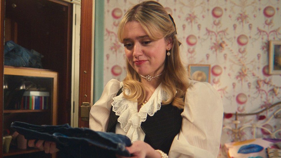 Aimee Lou Wood as Aimee in Sex Education. Aimee, a young white woman, has long blonde hair styled half-up-half-down with a short fringe to the side of her face. She wears a black waist coat over a frilly cream shirt and a pearl choker-style necklace. Aimee is pictured inside a bedroom with decorative pink and white wallpaper and holds a pair of jeans in front of her.