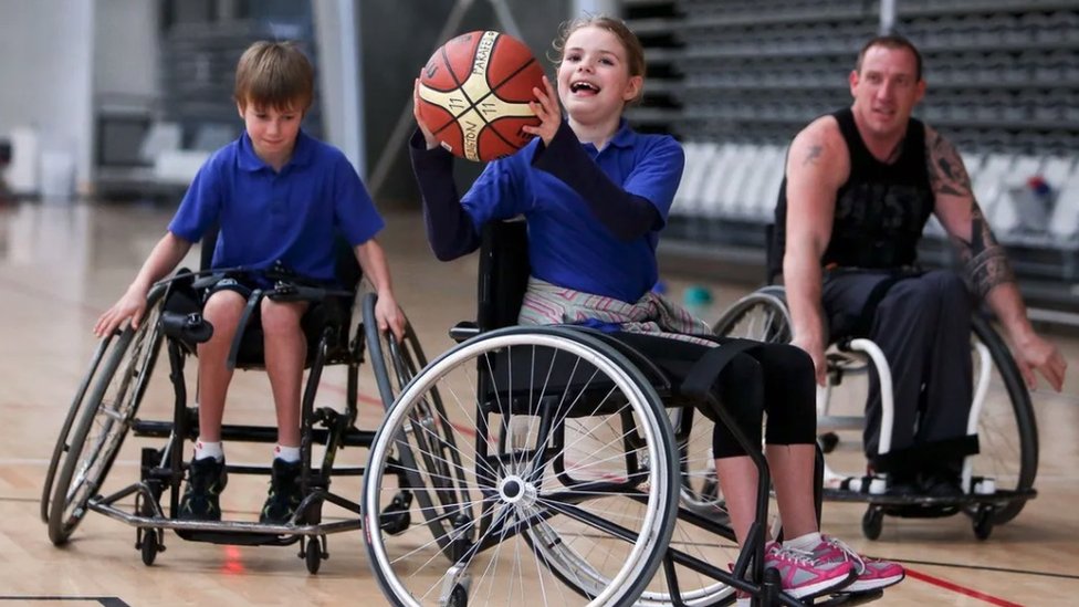 Two boys in wheelchairs play basketball while an adult also in a wheelchair coaches them