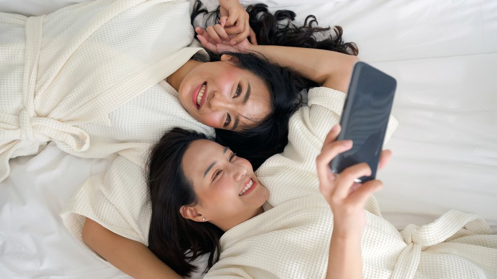 Two women lie on a bed and take a selfie