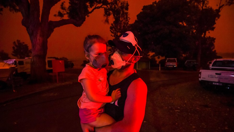Майк держит свою дочь Эллу, пока небо над головой становится красным днем ??4 января 2020 года в Маллакооте, Австралия