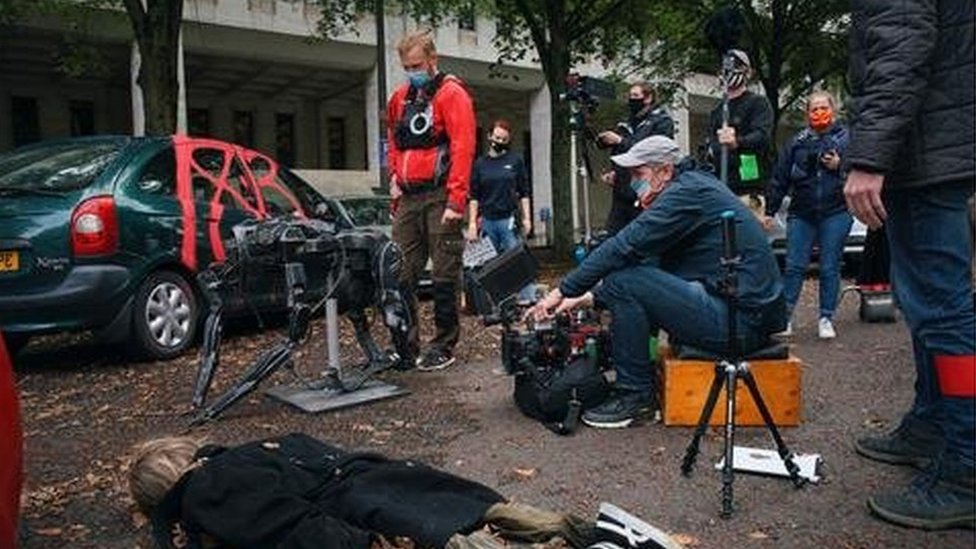 Актер лежит на полу в хаотической уличной сцене, оператор и команда в масках