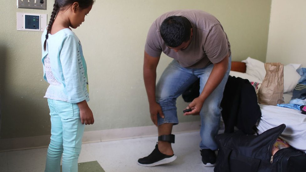 Una niña ve cómo su padre se pone una tobillera electrónica, requisito de las autoridades migratorias