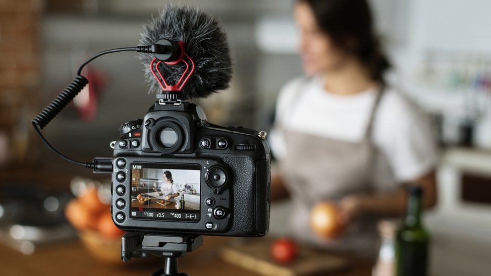 Una cocinera gravándose con una cámara mientras prepara un plato en su cocina.