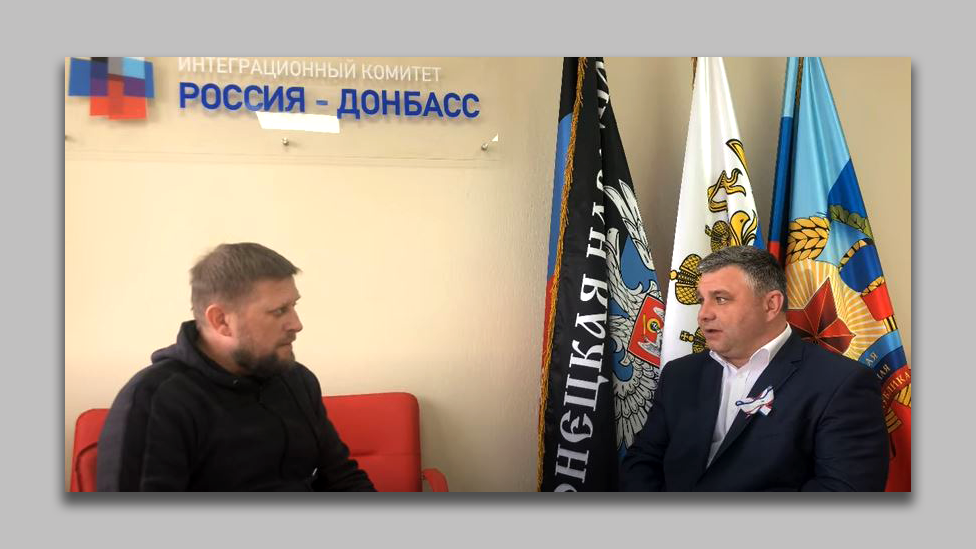 The logo of the Russia-Donbas Integration Committee visible on the wall above two men conducting an interview