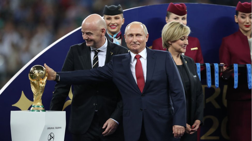 Putin sorrindo e tocando na taça, ao lado de Infantino, que ri também; atrás, aparecem mulheres que participam da organização do evento