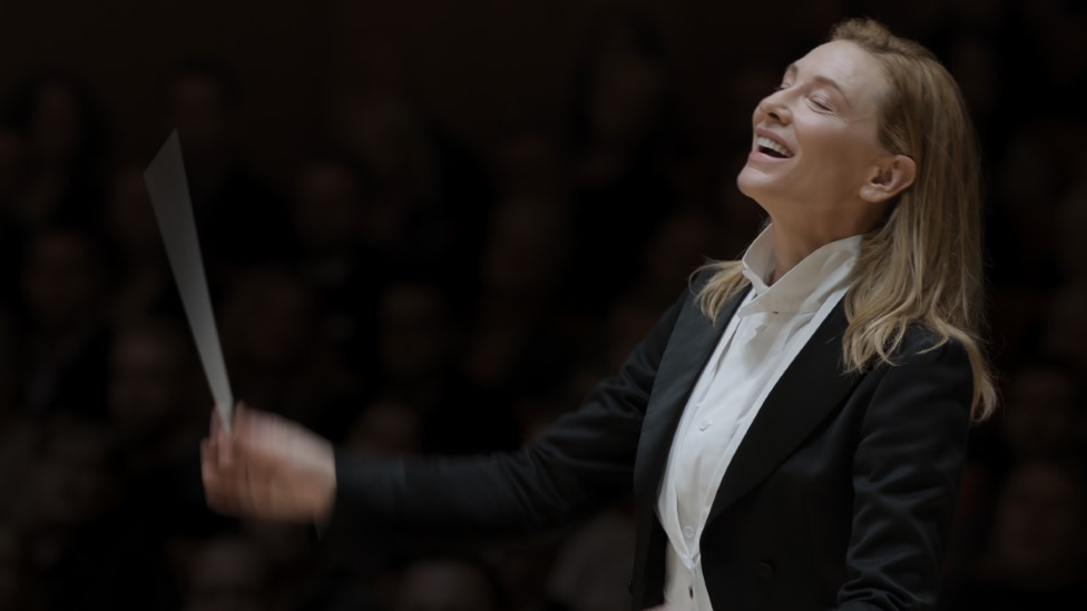 Cate Blanchett, uma mulher branca e loira, segurando uma batuta de maestro (ela aparente estar regendo uma orquestra)