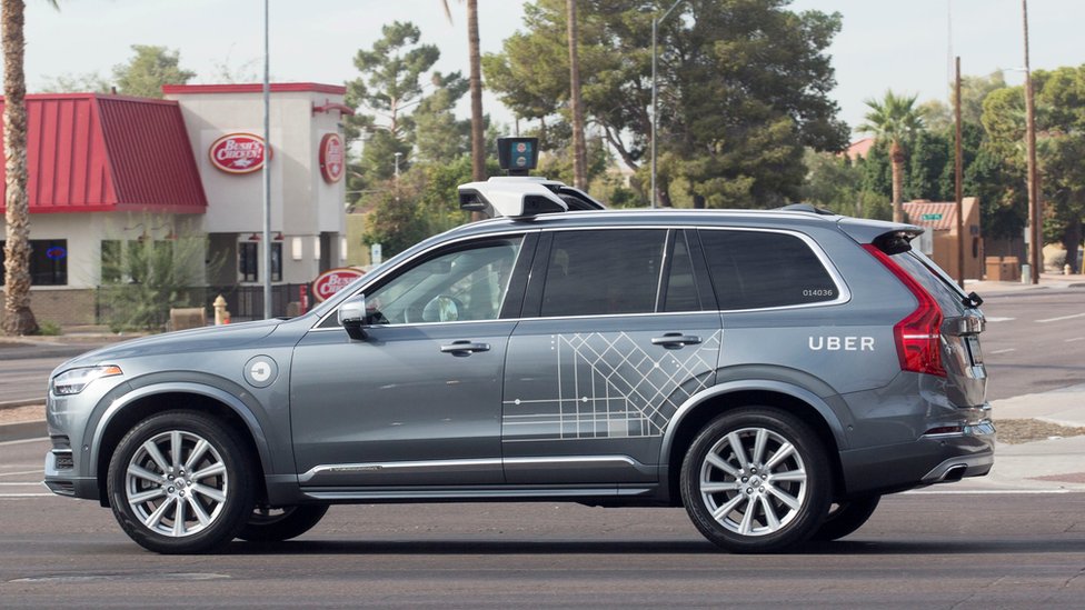 A metallic grey Volvo car, wrapped with some occasional Uber branding in white vinyl, is seen here with a large mount on top of the vehicle which houses self-driving equipment