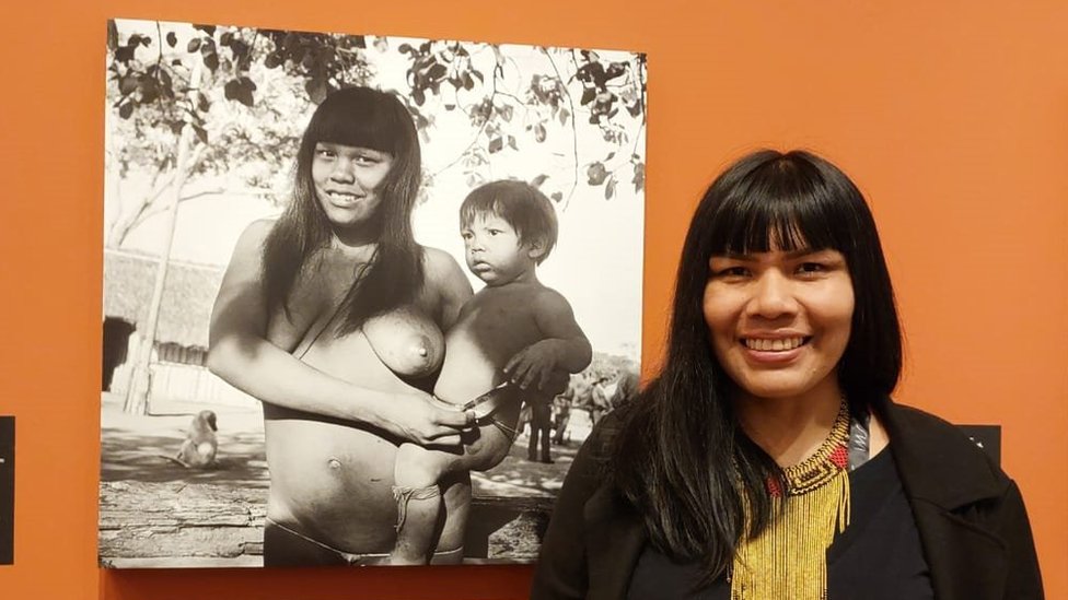 Fotografia colorida mostra uma mulher indígena de cabelo preto comprido e franja ao lado de uma fotografia preto e branca na parede na qual uma mulher indígena segura um bebê no colo
