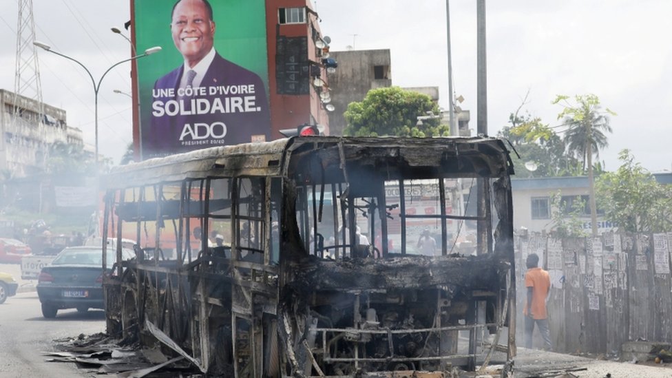 Рекламный щит президента Кот-д'Ивуара Алассана Уаттары за автобусом, сожженным протестующими против его решения баллотироваться на третий срок, в Абиджане, Кот-д'Ивуар - 19 октября 2020 г.