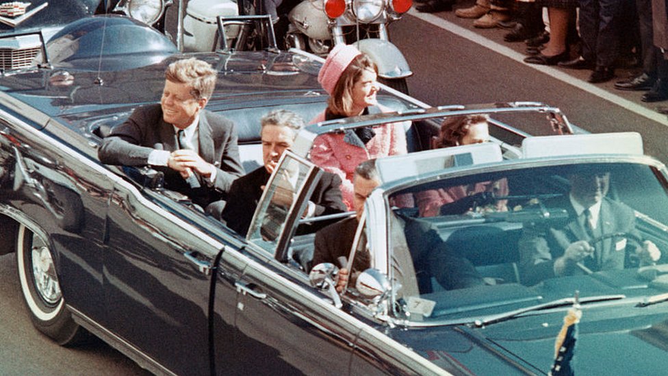 Kennedy y su esposa, Jacqueline, sonríen a al multitud desde un auto descapotado