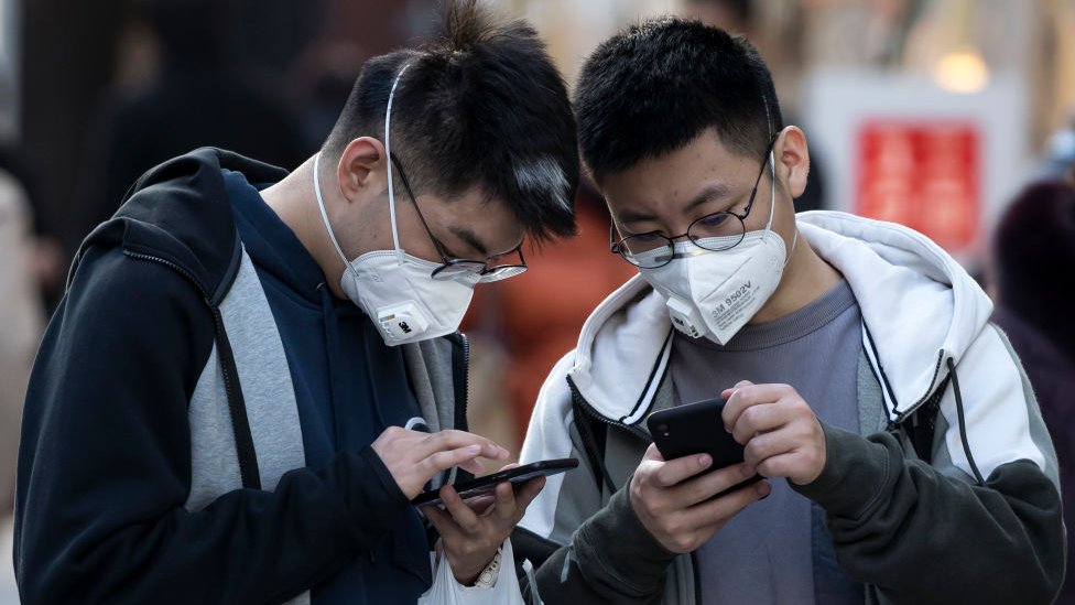 Dos jóvenes asiáticos con mascarilla revisan sus teléfonos.