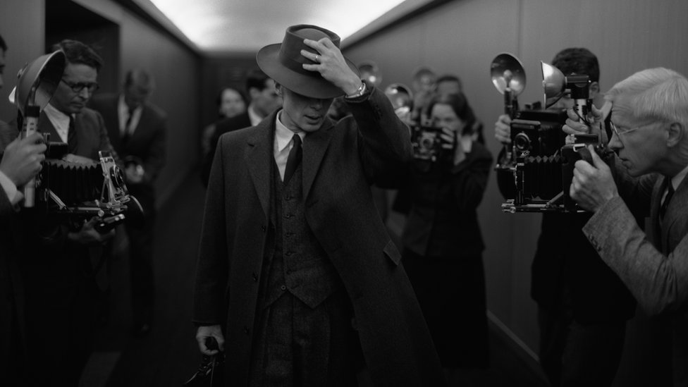 Fotografia em preto e branco mostra um homem branco de terno e chapéu cercado por fotográfos