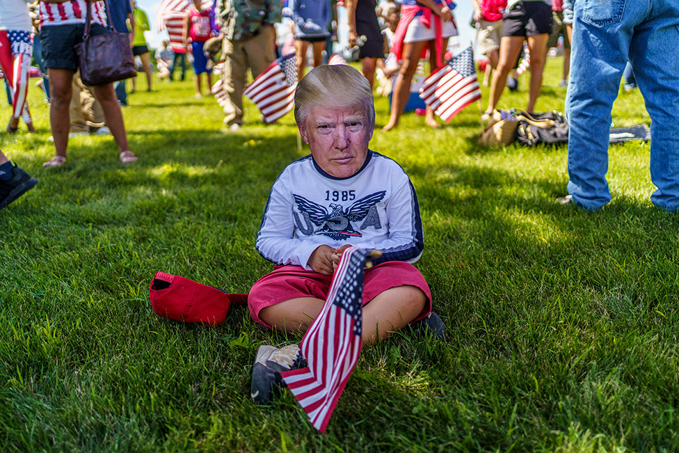 A supporter sits on the ground whilst wearing a mask of Donald Trump