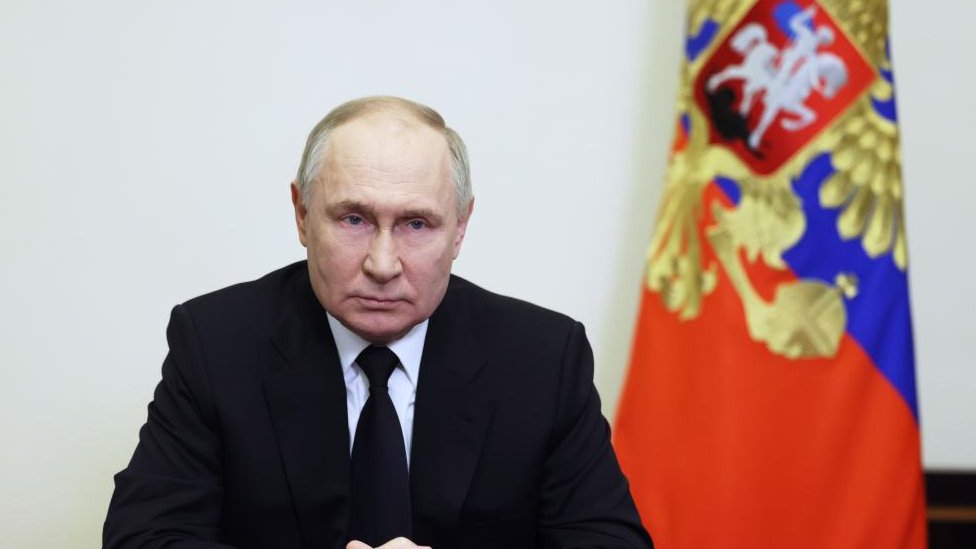 President Putin wearing a black suit and tie, sitting in front of a Russian flag