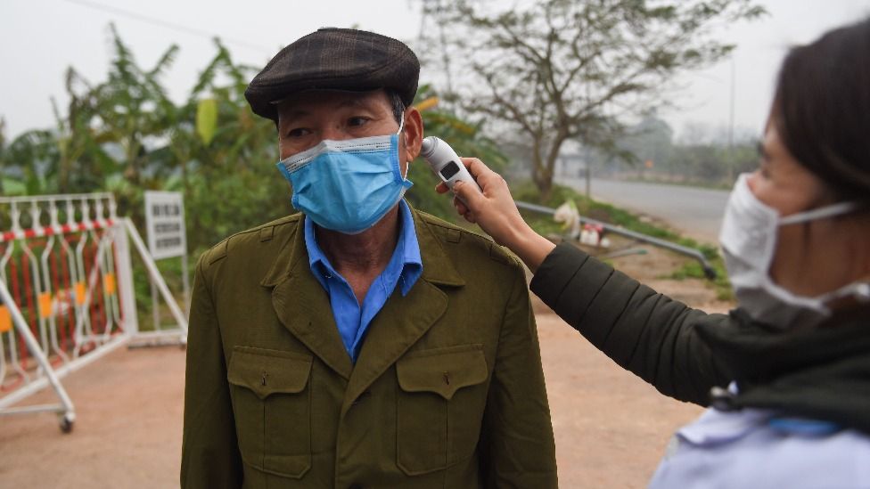 Seorang perempuan mengecek temperatur seorang laki-laki yang mengenakan masker.