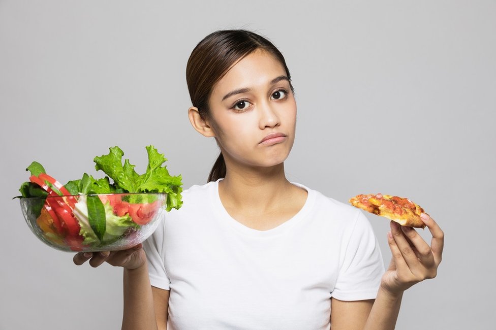 Una mujer joven sosteniendo una ensalada en una mano y en la otra un trozo de pizza.