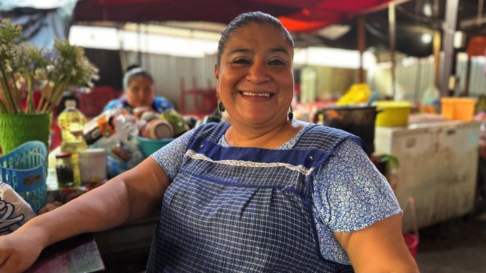 A large cheerful woman in front of a market stall selling food