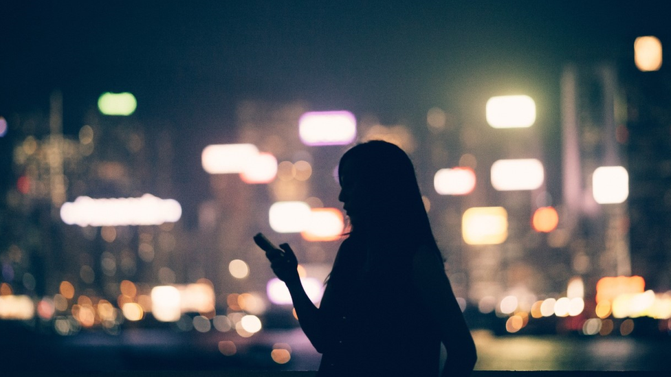 La sombra de una mujer mirando su télefono, sobre un fondo de luces de la calle.